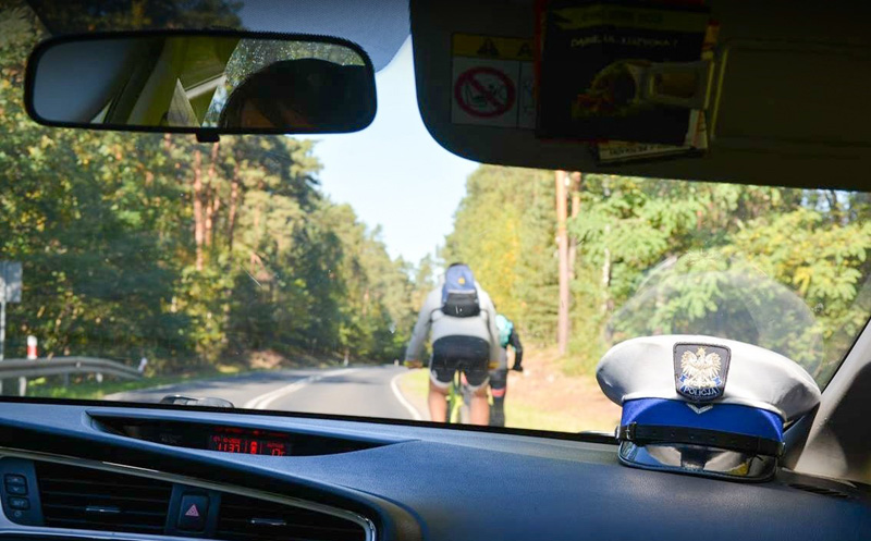 Rowerzyści widziani przez szybę radiowozu, w prawym dolnym rogu czapka policjanta ruchu drogowego.