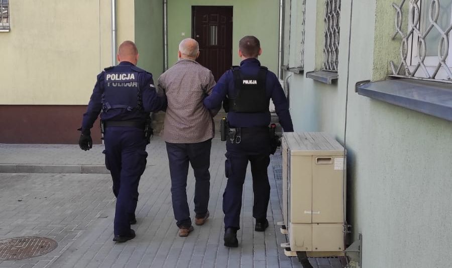 Dwaj umundurowani policjanci prowadzą zatrzymanego mężczyznę. Widok z tyłu.