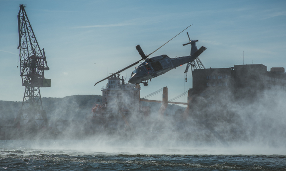 Śmigłowiec Black Hawk tuż nad wodą, za kurtyną drobnych kropek wody widać nabrzeże portowe.