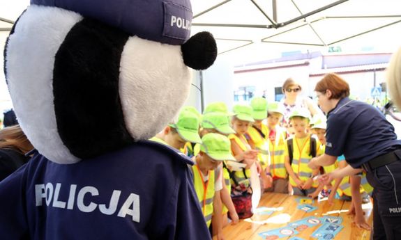 Policjantka rozdaje, dzieciom ubranym w kamizelki odblaskowe, ulotki na temat bezpieczeństwa, na pierwszym planie maskotka policyjna.