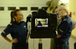 plan filmowy - na pierwszym planie aparat - w obiektywie widoczna Anna Mucha w policyjnym mundurze