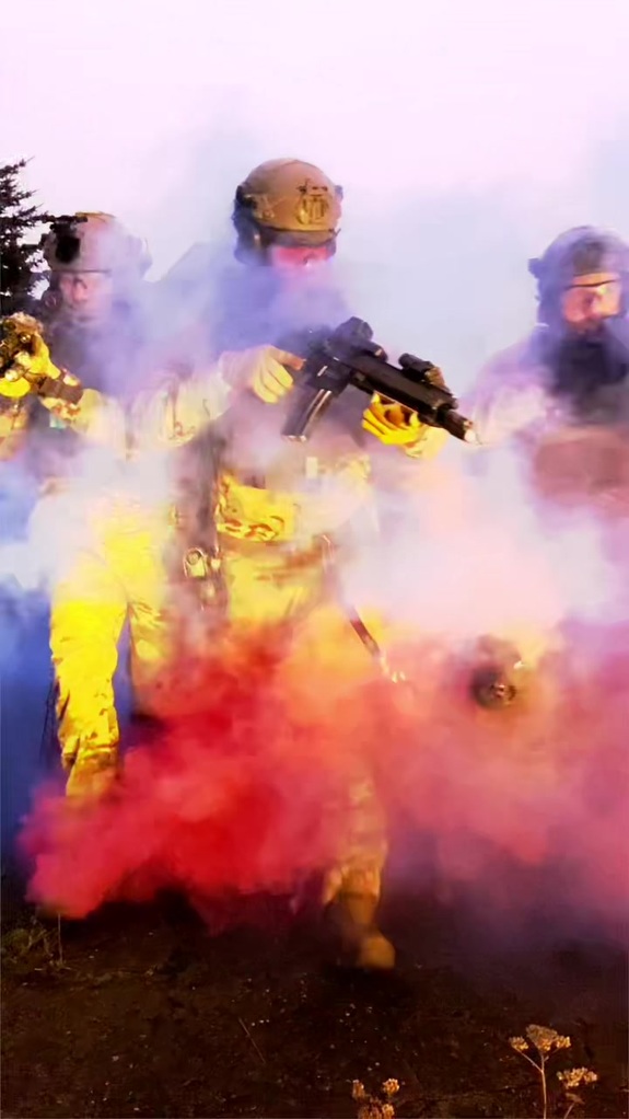 Trzej policjanci BOA wyłaniają się z wielokolorowego dymu z bronią gotową do strzału.