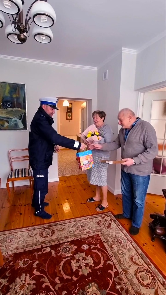 Policjant ruchu drogowego wręcza upominek mężczyźnie, obok stoi kobieta z kwiatami.