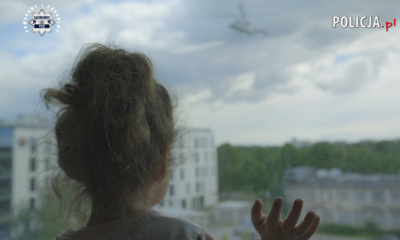 Dziewczynka obserwuje przez okno wskazuje policyjny helikopter widoczny na niebie.