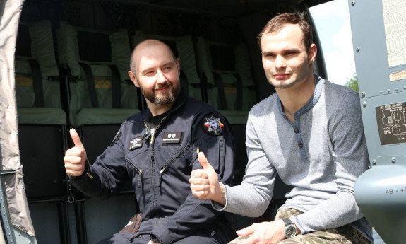 Policyjny pilot siedzi na skraju policyjnego śmigłowca z młodym mężczyzną. Obaj maja kciuki uniesione do góry.