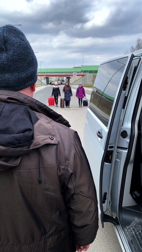 Mężczyzna stoi koło otwartych drzwi busa i obserwuje zbliżające się kobiety z walizkami.