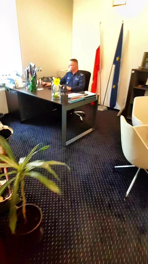 Policjant siedzi z biurkiem, w tle flagi Polski i Unii Europejskiej.