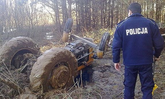 Policjant odwrócony tyłem patrzy w lewo na przewrócony do góry kołami żółty traktor