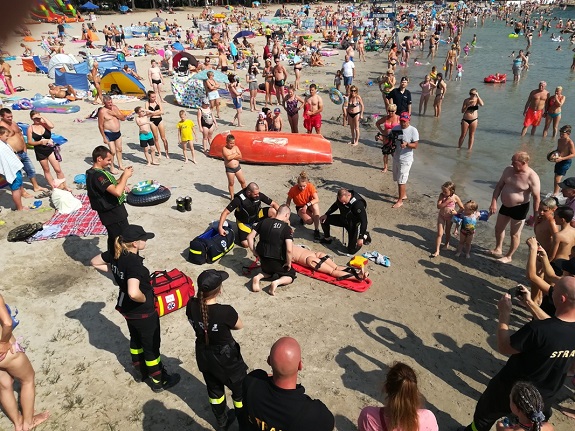 Policjanci i strażacy na plaży pokazują zasady udzielania pierwszej pomocy.