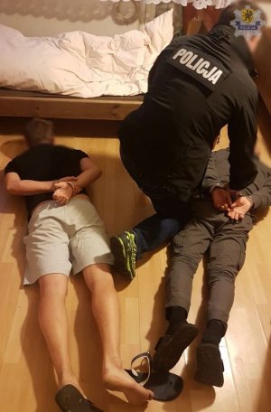 policjant z dwoma zatrzymanymi leżącymi na podłodze, jednemu z nich zakłada kajdanki