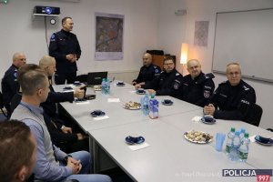 odprawa kadry kierowniczej, na zdjęciu salka konferencyjna, przy stole siedzą oficerowie policji, jeden oficer stoi i mówi do pozostałych, na ścianie powieszone dwie mapy i tablica