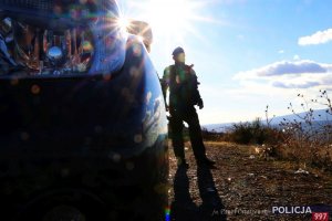 Na zdjęciu w ogromny przybliżeniu lampa samochodu, z tyłu w blasku słońca stoi policjant, ze względu na silne słoneczne światło postać ciemna