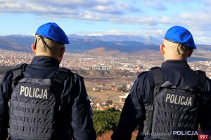 Dwaj policjanci stoją na wzgórzu, tyłem do aparatu, w głębi zdjęcia widać dolinę i miasto