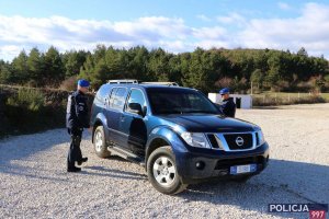 Dwaj policjanci w błękitnych beretach stoją obok radiowozu, gdzieś poza miastem