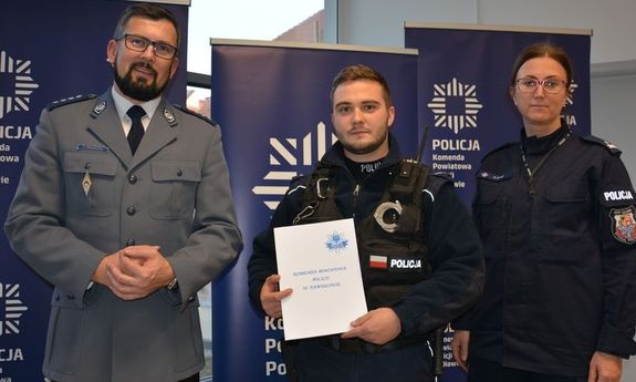 Na zdjęciu stoi troje policjantów, od lewej oficer, w środku wyróżniony policjant, po prawej kobieta oficer