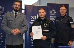 Na zdjęciu stoi troje policjantów, po lewej stronie oficer, w środku wyróżniony policjant, po prawej kobieta oficer