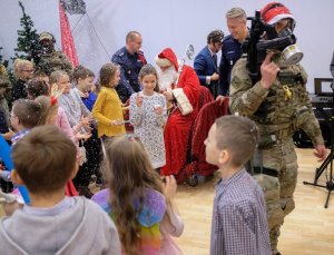 Dzieci stoją obok choinki obsypywane śniegiem. Obok dzieci św. Mikołaj. Na pierwszym planie policyjny komandos.