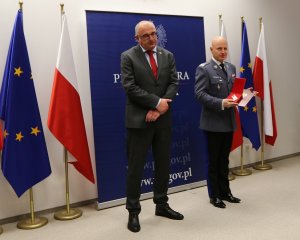 Na zdjęciu gen.insp. Jarosław Szymczyk Komendant Główny Policji prezentuje medal Zasłużony dla Prokuratury RP. obok stoi wyróżniony prokurator Michał Ostrowski – Dyrektor Departamentu do Spraw Przestępczości Zorganizowanej Prokuratury Krajowej. W tle flagi Polski i Unii Europejskiej.