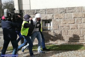 Zatrzymany prowadzony przez policjantów. Obecny także dziennikarz z kamerą.