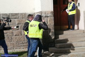 Policjanci prowadzą zatrzymanego. Zbliżają się do schodów prowadzących do budynku. Z tyłu podąża dziennikarz.