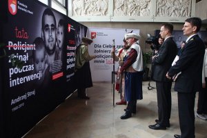 mężczyźni w garniturach i mężczyźni w strojach historycznych przed plakatem z napisem: Tata Policjant zginął podczas interwencji. Andrzej Struj1968-2010. Z tyłu operator kamery