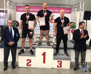 najlepsi zawodnicy stoją na podium, po obu stronach podium stoją dwaj mężczyźni