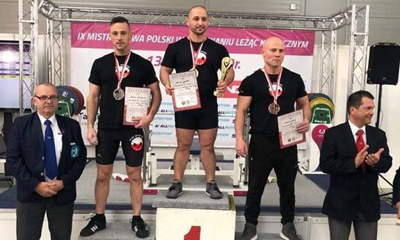 najlepsi zawodnicy stoją na podium, po obu stronach podium stoją dwaj mężczyźni