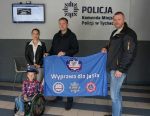 Jaś na wózku inwalidzkim w czapce policyjnej na głowie, za nim stoją dwaj policjanci trzymając  materiał z hasłem Wyprawa dla Jasia oraz loga organizacji, po lewej stronie stoi mama Jasia