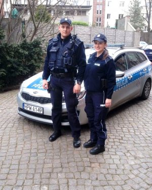 zdjęcie kolorowe na którym widać policjantkę i policjantka w granatowym mundurze na tle radiowozu oznakowanego