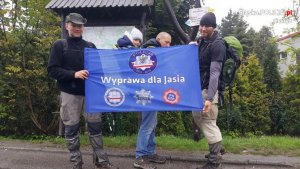 Uczestnicy wyprawy na mecie z flagą &quot;Wyprawa dla Jasia&quot;, z tyłu mężczyzna na plecach trzyma chłopca
