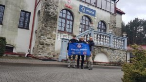 zdjęcie przedstawia trzech mężczyzn trzymających flagę z napisem Wyprawa dla Jasia na tle komendy Policji