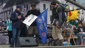 zdjęcie  przedstawia trzech mężczyzn na scenie, jeden z nich na plecach trzyma w foteliku chłopca, obok niego inny trzyma flagę z napisem Wyprawa dla Jasia i przyjmuje potwierdzenie zapłaty w postaci bonu od stojącego z lewej strony mężczyzny