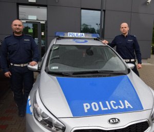 Mł. asp. Łukasz Pieszczek oraz sierż. Piotr Twardzik policjanci ruchu drogowego stoją przy oznakowanym radiowozie policyjnym