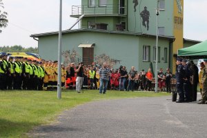 Na zdjęciu widać uczestników ćwiczeń terenowych, m.in. oddział policji oraz grupy poszukiwawczo ratownicze. Po prawej stronie stoją gospodarze i organizatorzy przedsięwzięcia, którzy rozpoczynają manewry witając wszystkich uczestników.  W tle budynek Grom Group.