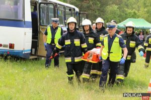 Na zdjęciu ratownik medyczny, kilku strażaków i policjant niosą na noszach ranną osobę. W tle widać autokar, który brał udział w wypadku.