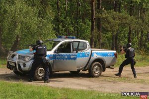 Na zdjęciu widać oznakowany samochód policyjny terenowy oraz dwóch funkcjonariuszy Policji w kamizelkach kuloodpornych i kaskach ochronnych na głowie z długą bronią. Stoją za radiowozem celując bronią w stronę niewidocznego na zdjęciu sprawcy porwania dziecka.