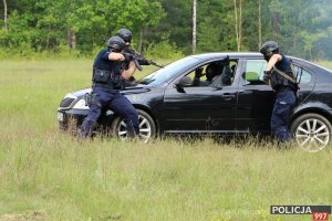 Na zdjęciu trzech funkcjonariuszy Policji w kominiarkach oraz kamizelkach kuloodpornych kierują broń w stronę sprawcy siedzącego na miejscu kierowcy w czarnym samochodzie osobowym.