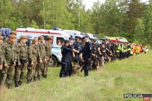 Na zdjęciu widać zgrupowanych w szeregu żołnierzy WOT, policjantów oraz członków grup poszukiwawczo ratowniczych. W tle samochody służbowe Policji, Straży Pożarnej oraz karetki.