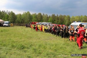 Na zdjęciu widać zgrupowanych w szeregu żołnierzy WOT, policjantów oraz członków grup poszukiwawczo ratowniczych. W tle samochody służbowe Policji, Straży Pożarnej oraz karetki.