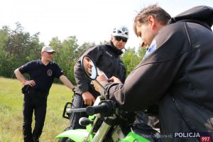 Na zdjęciu widać funkcjonariusza Policji – członka policyjnej nieetatowej grupy poszukiwawczo ratowniczej szykującego się do wyjazdu w teren na motocyklu terenowym. W tle pozostali członkowie grupy.