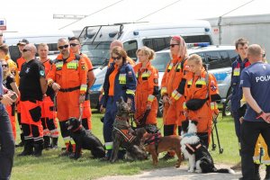 Na zdjęciu członkowie kilku grup poszukiwawczo ratowniczych z psami ratownicznymi. Wszyscy stoją w oczekiwaniu na wyznaczenie zadań.