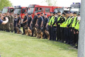 Na zdjęciu stoją funkcjonariusze Policji w zwartym szeregu. Za nimi stoi oddział Wojsk Obrony Terytorialnej. W tle samochody PSP.