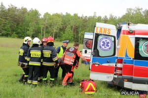 Na zdjęciu widać  jak ratownik medyczny, grupa strażaków oraz policjant umieszczają ranną osobę w karetce pogotowia. Obok nich stoi karetka z otwartymi drzwiami