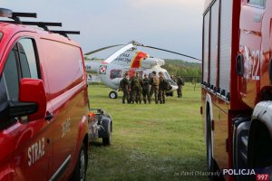 Na zdjęciu zbliżenie na dwa radiowozy strażackie, w oddali w tle śmigłowiec SG oraz grupa żołnierzy z 13 Batalionu Wojski Obrony Terytorialnej