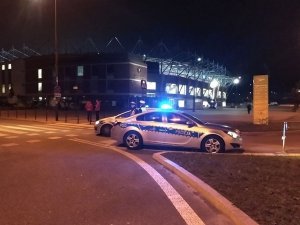 Na zdjęciu widoczne są dwa oznakowane radiowozy policji (Wydziału Ruchu Drogowego), które stoją zaparkowane w okolicach stadionu miejskiego w Łodzi przy ul. Piłsudskiego. Radiowozy biorą udział w zabezpieczeniu meczu. Zdjęcie wykonane w porze nocnej