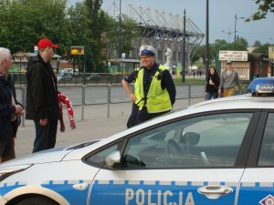 Na zdjęciu widoczni są policjanci Ruchu Drogowego KMP w Łodzi, podczas rozmowy z dwójką kibiców. Jedne z mężczyzn na ma głowie czerwoną czapkę i trzyma w ręku szalik reprezentacji Polski. Z tyłu widoczna para, kobieta mężczyzna, pas ulicy oraz stadion miejski