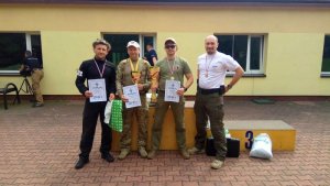 czterej zawodnicy stoją przy podium, trzech z nich trzyma dyplomy