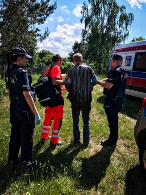 na środku zaginiony 66-letni mężczyzna. Po jego lewej stronie ratownik medyczny, po prawej umundurowany policjant. Po ich lewej stronie stojąca policjantka z założonymi jednorazowymi rękawiczkami. Po prawej stronie karetka pogotowia