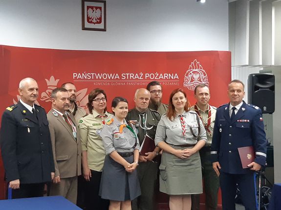 Umundurowani Pierwszy Zastępca Komendanta Głównego Policji, Komendant Główny Państwowej Straży Pożarnej, Dyrektor Lasów Państwowych wraz z harcerzami na tle banneru Państwowej Straży Pożarnej.