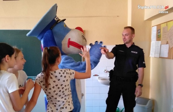 Policjant w trakcie spotkania z dziećmi. W tle widoczna maskotka policyjna.